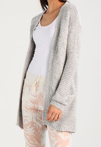 Cardigan lavorato a maglia grigio chiaro con apertura frontale, maniche lunghe e due tasche anteriori. Tessuto testurizzato e dettaglio a costine sull'orlo. Abbinato a pantaloni con motivo.