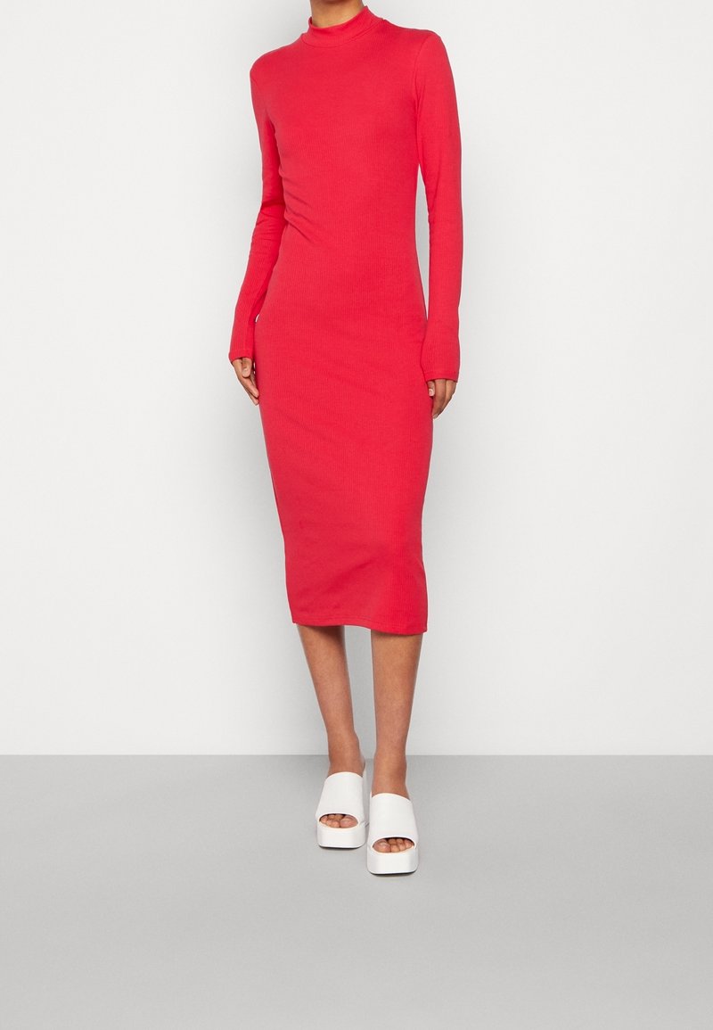 Femme portant une robe rouge ajustée à manches longues, arrivant au genou, et des sandales compensées blanches, se tenant devant un fond clair et uni.
