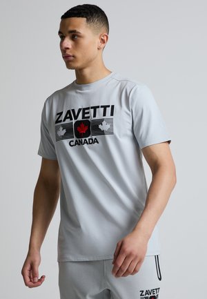 Ung mand iført lysgrå Zavetti Canada t-shirt og matchende shorts, står mod en ensfarvet grå baggrund og kigger til venstre.