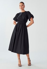 Črna midi obleka s pufastimi rokavi in elastičnim pasom. Gladek material in sproščen kroj, v kombinaciji s črnimi sandalami na vezi.