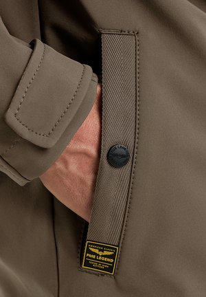 Hand in einer braunen Jackentasche mit einem Stoffstreifen mit schwarzem Knopf und einem gelb-schwarzen PME Legend Label.