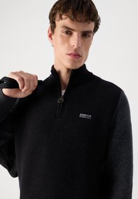 Sweater meio zip em preto e cinza, feito de um tecido suave e texturizado. Apresenta um logótipo no peito e punhos canelados. Adequado para uso casual.
