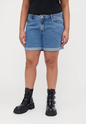 Persoon die opgerolde blauwe denim shorts en zwarte leren enkellaarzen met gespen draagt, staand tegen een effen achtergrond.