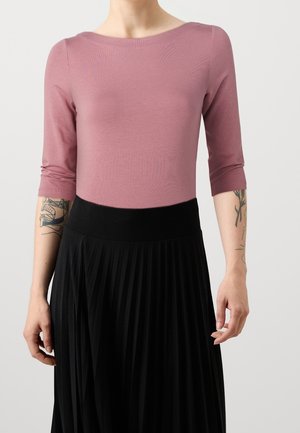 Osoba ubrana w bluzkę o rękawach długości do łokcia w kolorze fioletowo-szarawym i wysoką, plisowaną czarną spódnicę, z widocznymi tatuażami na obu przedramionach.