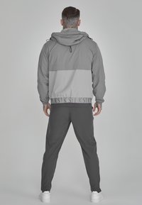 Siva zip-up jakna z lažjo sivo ploščo po sredini, v kombinaciji s temnosivimi hlačami in belimi supergami. Ima nastavljivo kapuco.