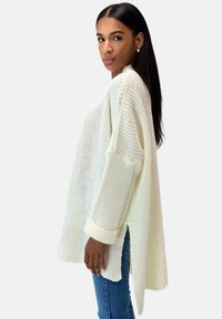 Frau mit langen dunklen Haaren, die einen lockeren, weißen Strickpullover mit seitlichen Schlitzen und blaue Jeans trägt, steht vor einem schlichten Hintergrund.