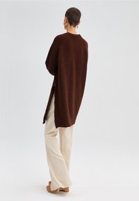 Langer, brauner Strickcardigan mit gerippter Textur, der eine lockere Passform und seitliche Schlitze aufweist. Kombiniert mit crema-farbenen, weiten Hosen und Plateauschuhen.