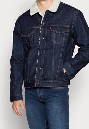Mann trägt eine dunkelblaue Jeansjacke mit weißem Fleece-Kragen und passende dunkelblaue Jeans, eine Hand in der Tasche.
