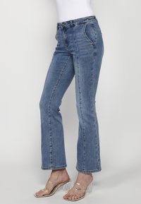 Blå denimbyxor med vida ben och hög midja, framfickor och sömmar som detalj. Bärs med klara klacksandaler. Slät textur.