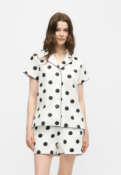 Femme portant une chemise blanche à manches courtes boutonnée et un short assorti avec de grands pois noirs et des détails en passepoil noir.