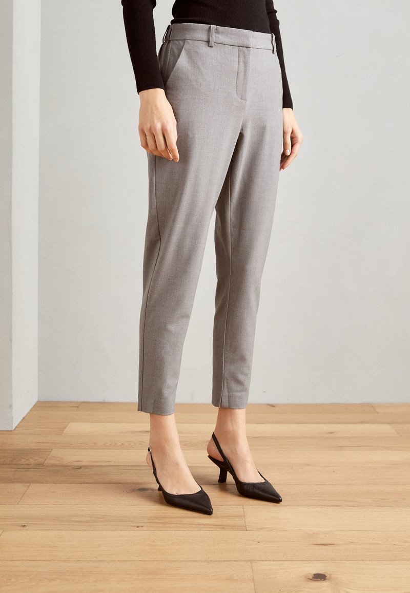 Pantalon gris sur mesure, coupe slim, avec poches latérales et ceinture plate, assorti à des escarpins noirs à bouts pointus et bride arrière.