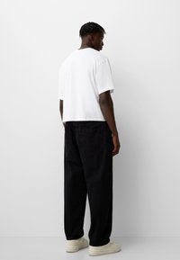 T-shirt bianco in cotone con maniche corte, abbinato a pantaloni in denim neri larghi. Le scarpe presentano un colore neutro chiaro e un design minimalista.