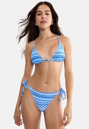 Kvinde iført en blå og hvid bikinitop med zigzag-mønster og bindebånd i siden, stående mod en ensfarvet lys baggrund.