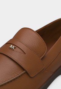 Καφετί μοκασίνι από δερμάτινη υφή με λουρί που διαθέτει μικρή ασημένια λεπτομέρεια λογότυπου "MK" σε ομαλή λευκή επιφάνεια.