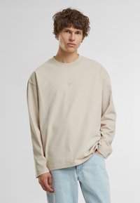 Beige Langarmshirt mit Rundhalsausschnitt und bequemer Passform. Hat ein dezentes Logo auf der Brust und besteht aus weichem Baumwollmaterial.