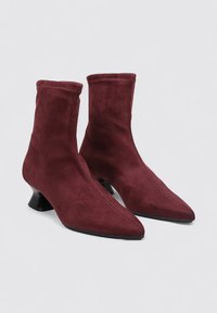 Botas de ante burdeos hasta el tobillo con punta afilada, parte superior elástica y un tacón negro curvado. Superficie texturizada y silueta elegante.