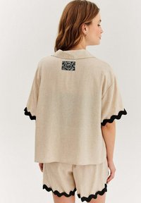 Béžová lněná košile s krátkým rukávem a černým scalloped lemem a černou vzorovanou nášivkou na zádech, doplněná o odpovídající šortky.