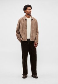 Brūngana zamšādas jaka, krēmkrāsas t-krekls un tumši brūnas kordura bikses. Apģērbā ir rāvējslēdzēja aizdare un sānu kabatas ar teksturētām audumu virsmām.