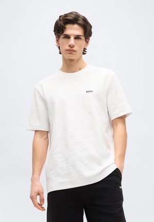 Mladi moški z valovitimi rjavimi lasmi, oblečen v belo teksturirano BOSS majico in črne kratke hlače, stoji pred enakomerno svetlo ozadje.