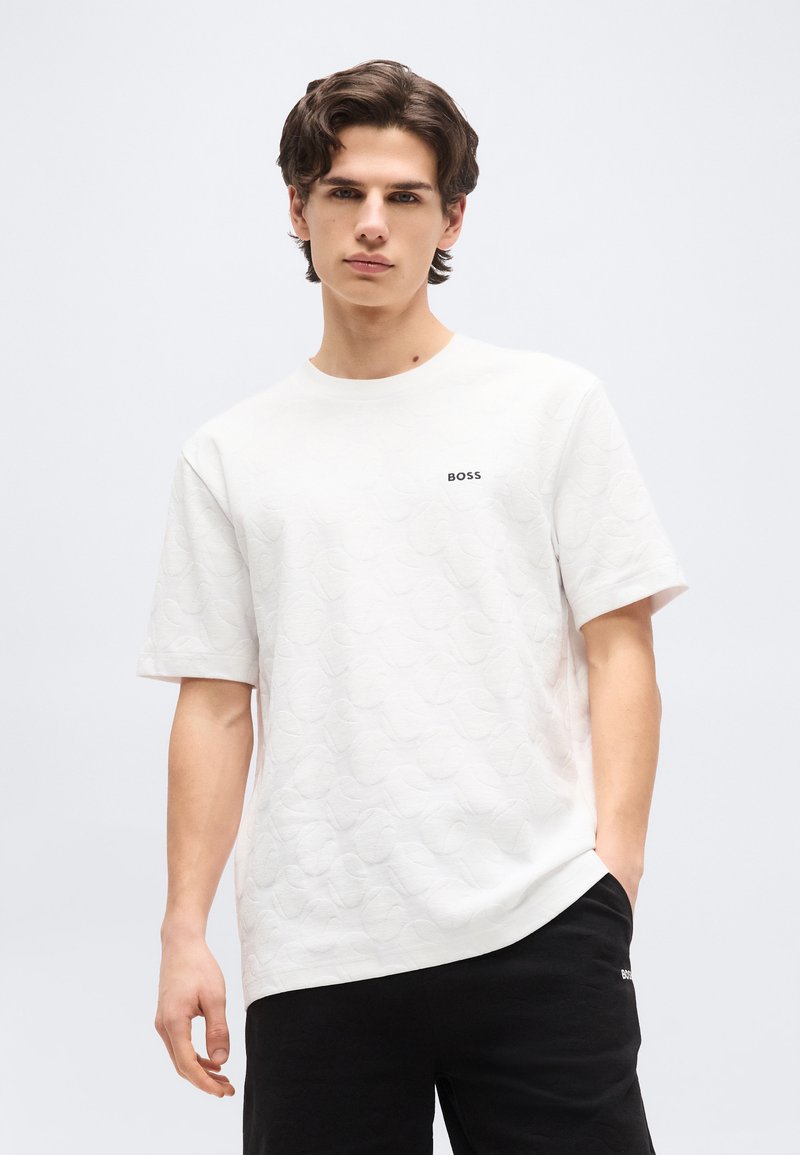 Νεαρός άνδρας με κυματιστά καστανά μαλλιά, φορώντας ένα λευκό υφασμάτινο μπλουζάκι BOSS και μαύρο σορτς, στέκεται μπροστά σε ένα απλό ανοιχτόχρωμο φόντο.