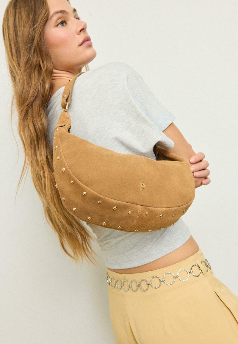Jeune femme aux longs cheveux portant un haut gris et un pantalon beige, avec un sac croissant en daim beige orné de clous dorés sur l'épaule.