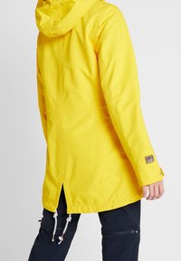 Jasnożółty wodoodporny płaszcz z kapturem noszony na czarnych spodniach, widok z tyłu z regulowanymi sznurkami ściągającymi oraz drobnym detalem na nadgarstku.