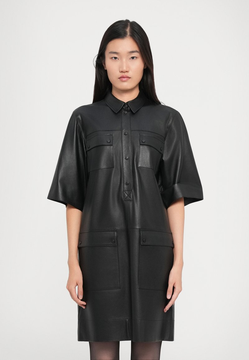 Γυναίκα φορεμένη με μαύρο δερμάτινο φόρεμα πουκαμίσα με κοντά μανίκια, κουμπωτό μπροστά και τέσσερις τσέπες με καπάκι, στέκεται μπροστά σε λευκό φόντο.