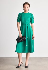 Πράσινο φόρεμα μέχρι το γόνατο με κοντά φουσκωτά μανίκια και ζώνη που δένει στη μέση. Μαύρη τσάντα-clutch και μαύρες γόβες με尖尖 το σχήμα συμπληρώνουν την εμφάνιση.