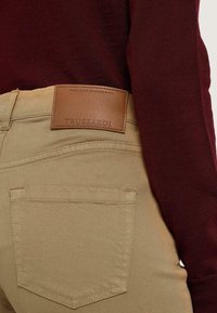 Pantalones de algodón beige con bolsillos traseros, que presentan una etiqueta de cuero marrón grabada con "TRUSSARDI". Suéter burdeos visible en la parte superior.