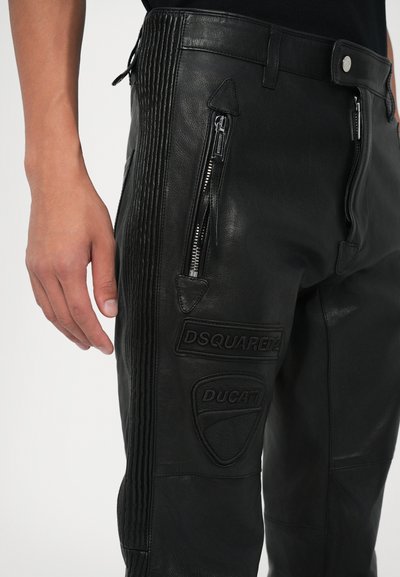 Ruka muža vedľa čiernych kožených nohavíc so zipsovým vreckom a prešívanými nášivkami DSQUARED2 a DUCATI na stehnách.