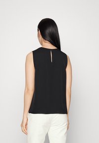 Schwarzes ärmelloses Top mit rundem Ausschnitt und einem Schlüsselaugen-Detail auf der Rückseite. Glattes Material, figurbetonter Schnitt, kombiniert mit weißen Hosen.