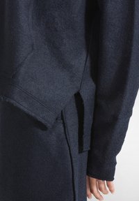 Mörkblå ulltröja med texturerad yta. Har överdimensionerade ärmar och en sidoklyv för flexibilitet. Synliga sömnadsdetaljer.