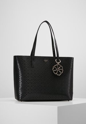 Sac fourre-tout noir Guess avec motif de logo embossé, doubles poignées et pendentif trèfle à quatre feuilles suspendu sur un piédestal blanc.