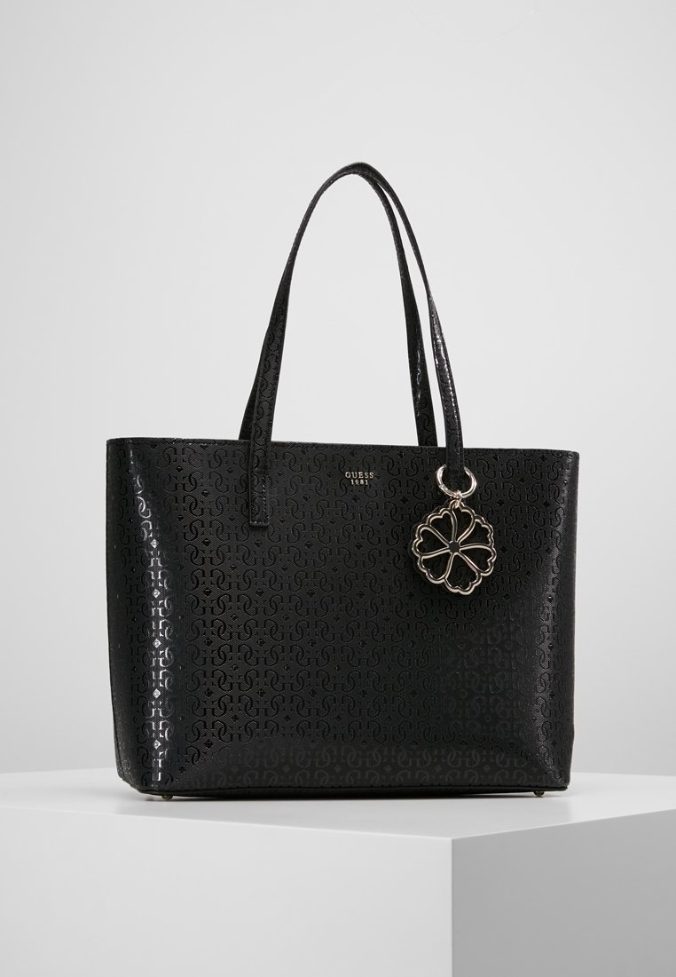 Sac fourre-tout noir Guess avec motif de logo embossé, doubles poignées et pendentif trèfle à quatre feuilles suspendu sur un piédestal blanc.