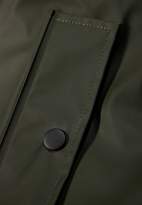Tela impermeable color verde oliva con una textura suave, que presenta un botón negro en contraste y costuras en contraste en un bolsillo de parche.
