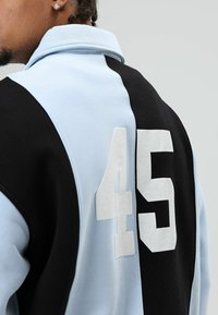 Sweat-shirt bicolore noir et bleu clair avec un col à revers. Présente un numéro "45" texturé en blanc dans le dos, fabriqué en matériau doux.