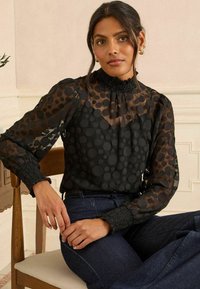 Blusa nera in tessuto trasparente, con motivo a pois, colletto alto a balze e polsini arricciati. Abbinata a jeans scuri.