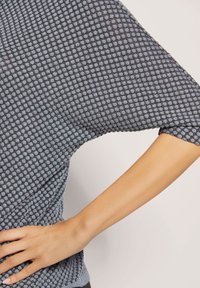 Blauwe en grijze gemêleerde trui met een ontspannen pasvorm, voorzien van een honingraatpatroon en brede mouwen. Hand rustend op heup.