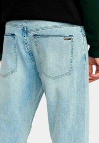 Vaqueros de mezclilla azul claro con bolsillos traseros, uno de ellos con una pequeña etiqueta negra y naranja de Volcom, combinados con una blusa negra y verde oscuro.