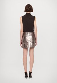 Femme aux cheveux courts et bouclés porte un haut noir sans manches, une mini-jupe argentée métallique et des sandales noires à talons hauts, debout de dos.
