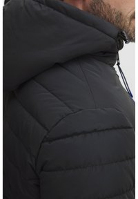 Svart polstret jakke med hette, vertikal kanaler og justerbar snor. Materialet har en glatt, litt glitrende tekstur.
