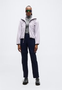 Γυναίκα που φορά γυαλιά ηλίου αθλητικά με ανακλαστικούς φακούς, ανοιχτό μοβ μπουφάν με φερμουάρ, γκρι μπλούζα με ψηλό γιακά, μπλε σκούρο παντελόνι και παπούτσια πεζοπορίας, στέκεται σε λευκό φόντο.