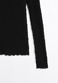 Black long-sleeve top made of textured fabric with a woven pattern. The hem is scalloped, adding a detail to the overall design.