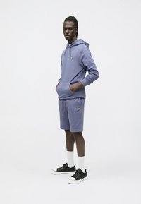 Vijoličasta majica s kapuco iz bombažne mešanice s sprednjim žepom, v kombinaciji z ujemajočimi kratkimi hlačami. Ima majhen rumen logotip. Črni supergi in bele nogavice.