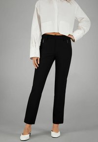 Schwarze, maßgeschneiderte Hose mit vorderen Reißverschlusstaschen, kombiniert mit einer verkürzten weißen Bluse mit Knöpfen. Die Schuhe sind weiß und haben einen niedrigen Absatz.