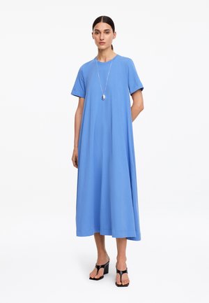 Donna che indossa un abito midi azzurro chiaro a maniche corte e ampio, con una collana con ciondolo in argento e sandali neri con tacco.