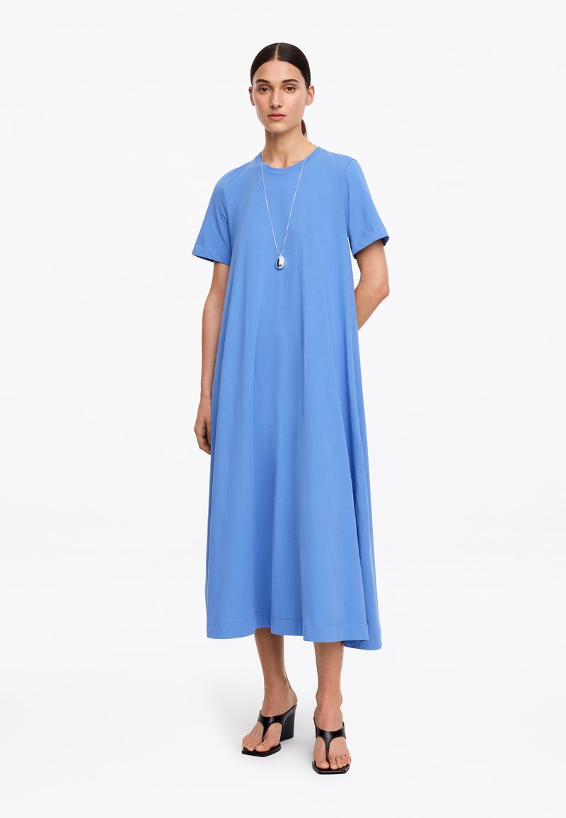 Femme portant une robe midi ample bleu clair à manches courtes, un collier pendentif en argent et des sandales noires à talons.