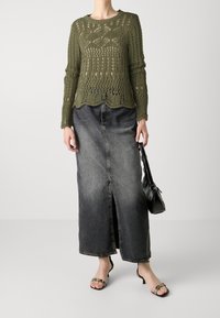 Pull en maille vert olive avec un design ajouré élaboré, associé à une jupe longue en denim noir avec une fente devant et un sac noir.