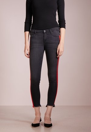 Personne portant un jean skinny noir raccourci avec des bandes latérales rouges et des ourlets effilochés, associé à un haut noir à manches longues et des chaussures noires pointues.