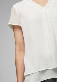 Blouse blanche à manches courtes en tissu léger, avec un col en V et des détails plissés aux épaules. Texture lisse, coupe décontractée.